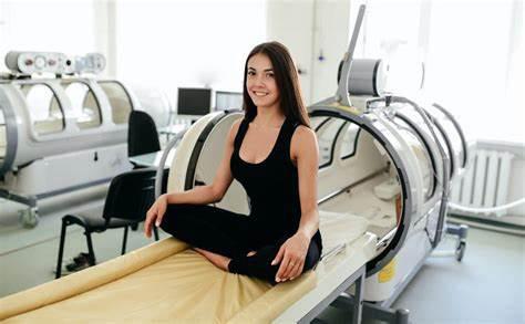 A person in athletic attire sits cross-legged on a hyperbaric chamber bed in a clinical setting, with medical equipment in the background.