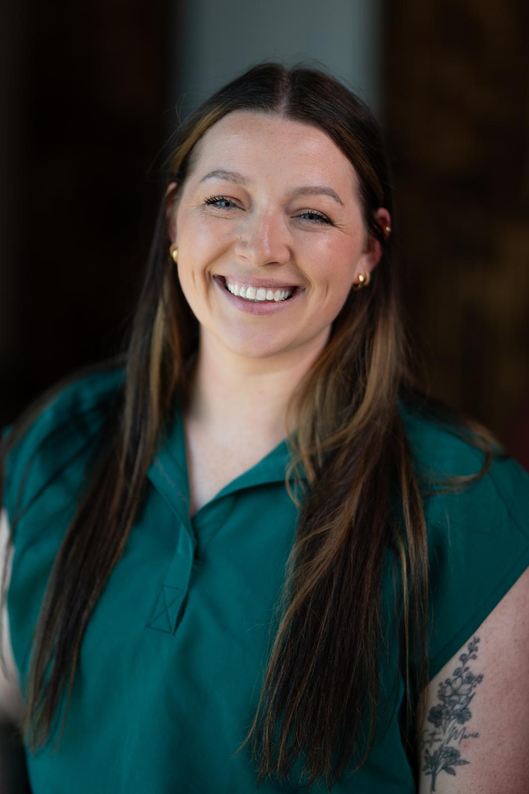 A person with long hair and a tattoo smiles warmly while wearing a teal shirt. The background is softly blurred, emphasizing their expression.