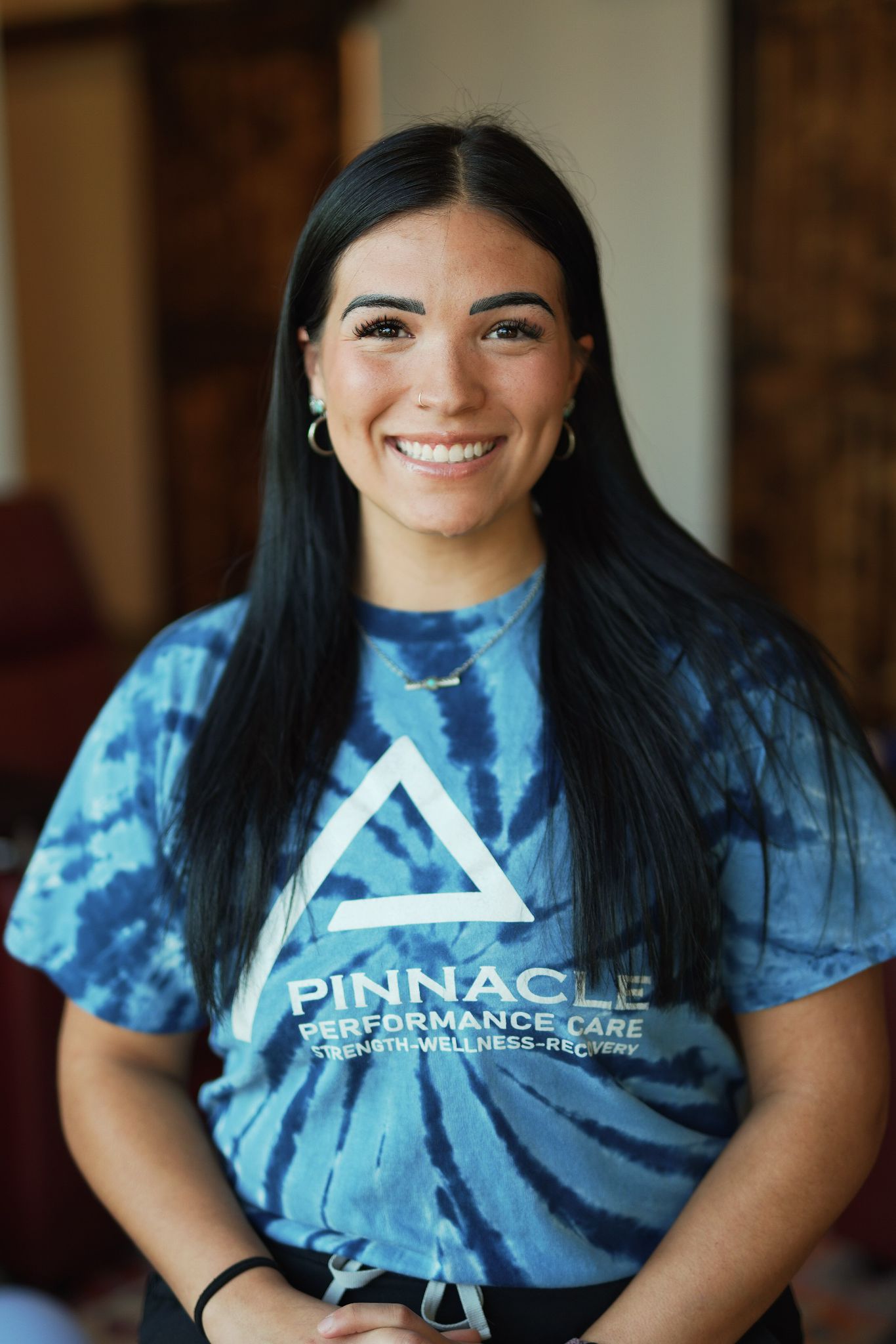 A person in a blue tie-dye shirt with a triangle logo smiles warmly inside a cozy, blurred indoor setting.